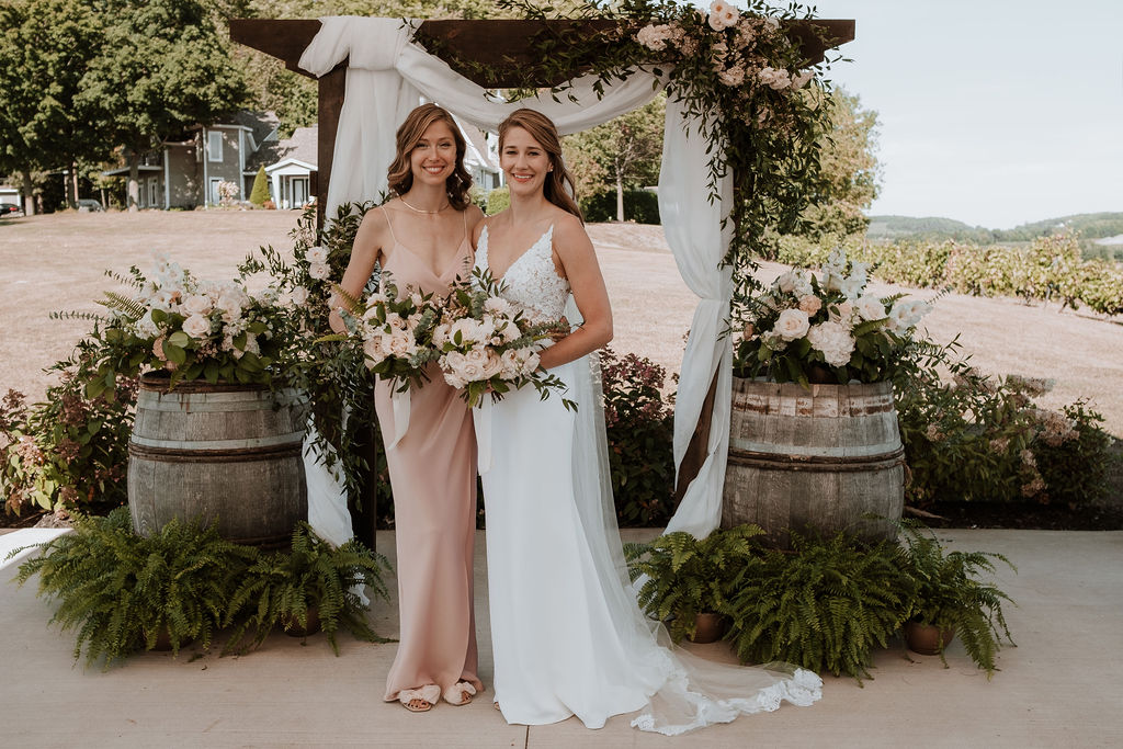 Bride and bridesmaid in blush dress with bouquets in front of ceremony arch
