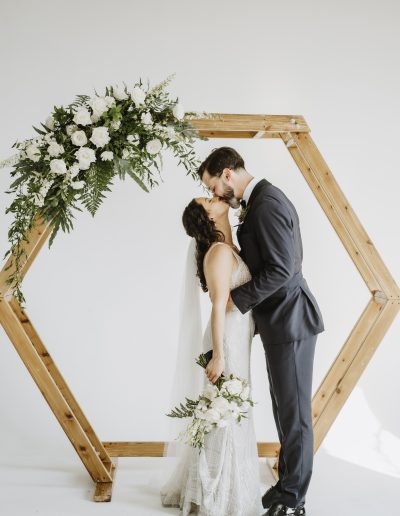 Bride and groom kissing in front of wood arch with white and green flowers | Flourish & Knot