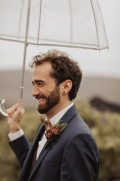 Groom under clear umbrella in navy suit with burgundy boutonniere