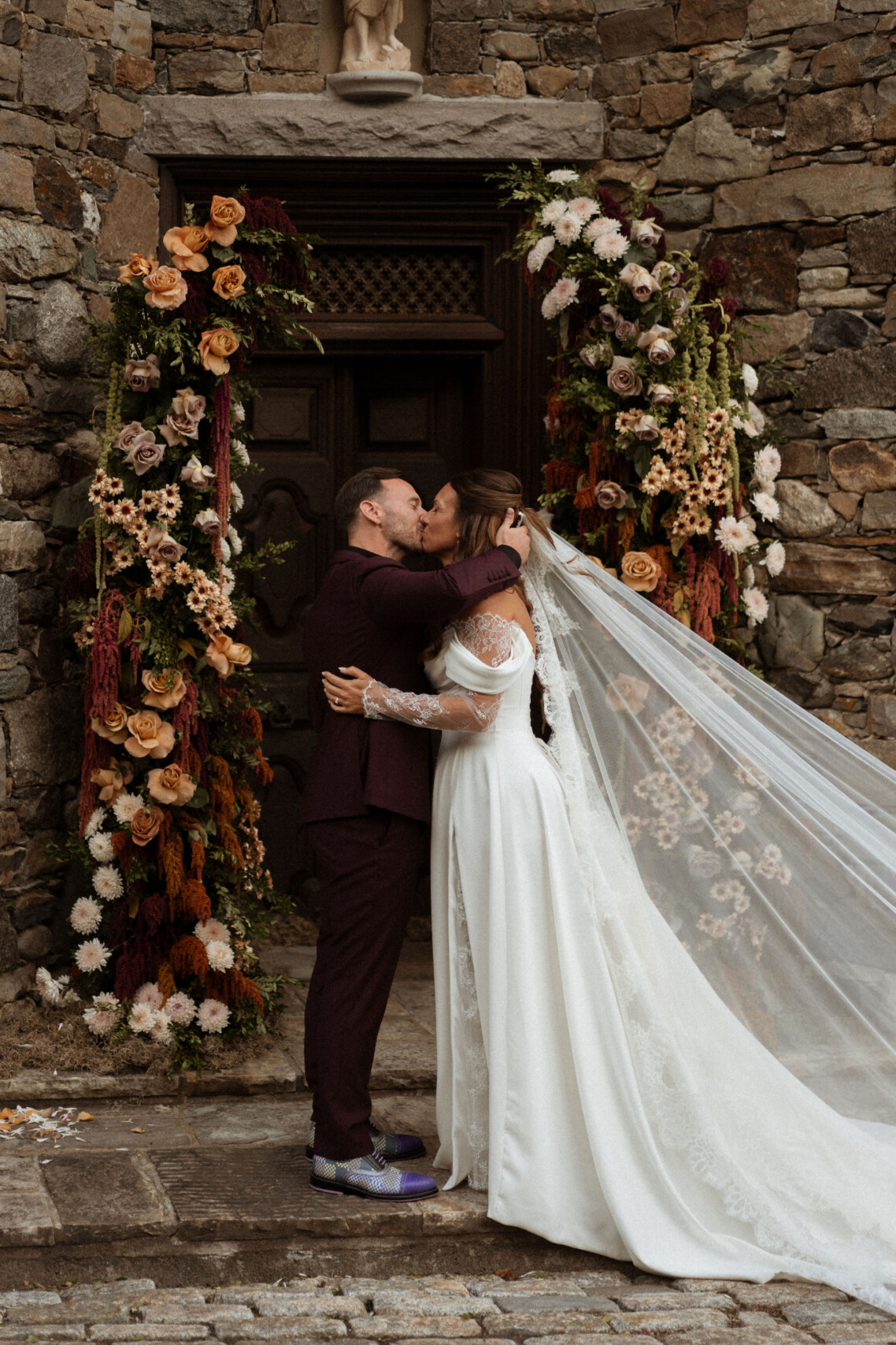 Bride and groom kissing under an abundant autumn wedding floral arch at the Château Ste-Agnès stone chapel, designed by Montreal wedding planner and florist Flourish and Knot.