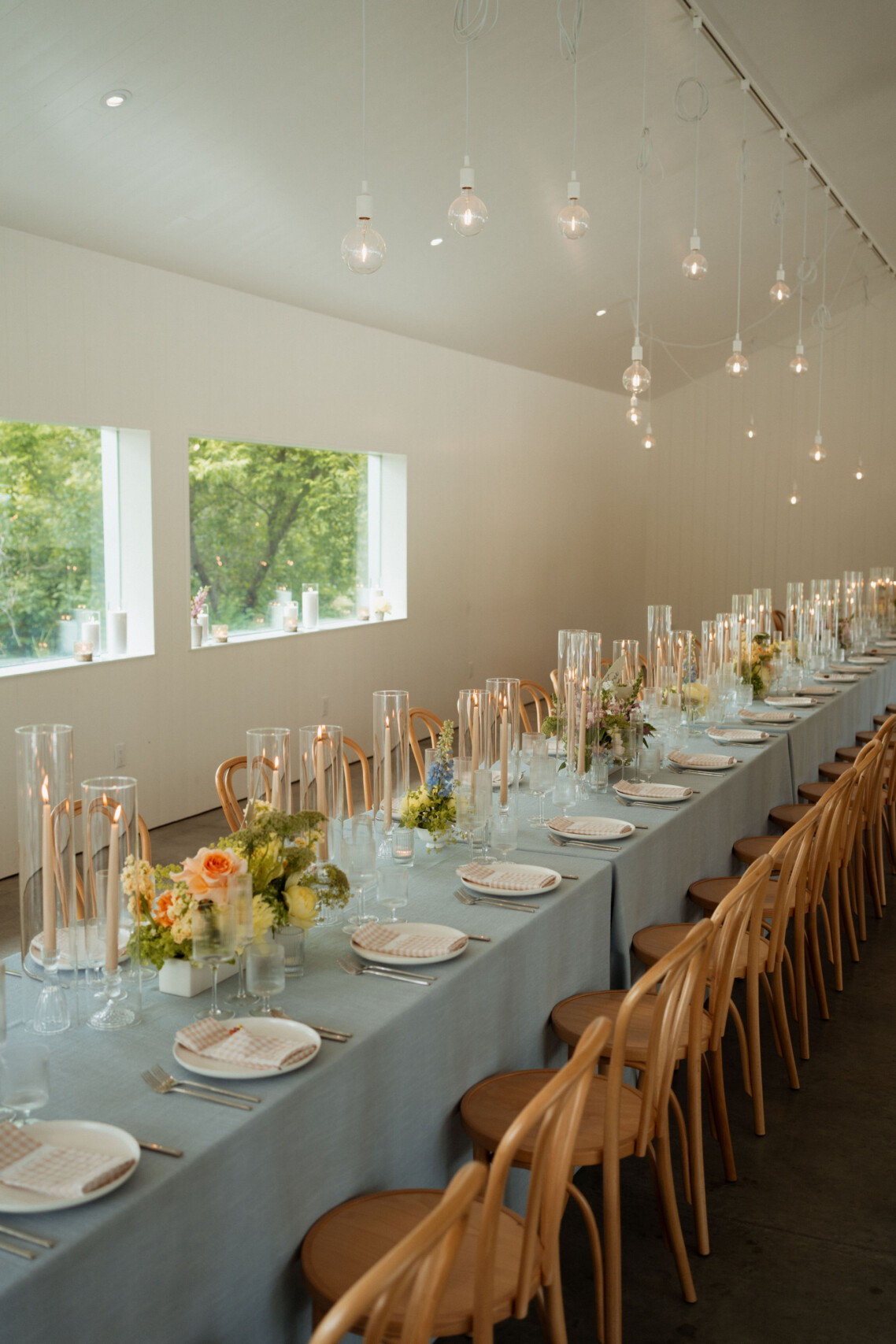 A high-angle vertical view of the minimalist wedding reception hall at Le Peaches & Cream, showcasing an ultra-long banquet table styled with dusty blue linens, modern hanging bulb fixtures, and a large window looking out onto lush trees.